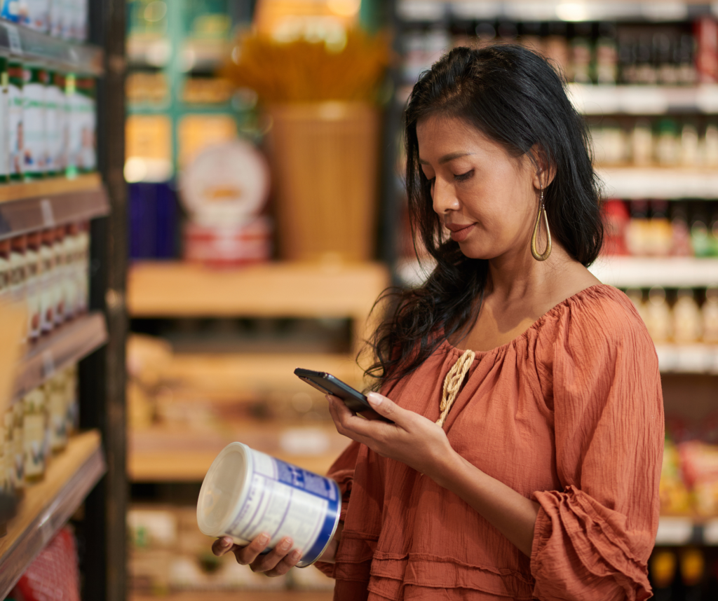 A lady in an orange top in a store, holding a product she's interested in and checking information online